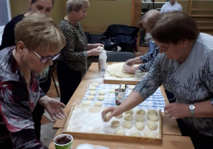 Seniorzy przygotowują pączki wykonane z ciasta i układają je na tacach.